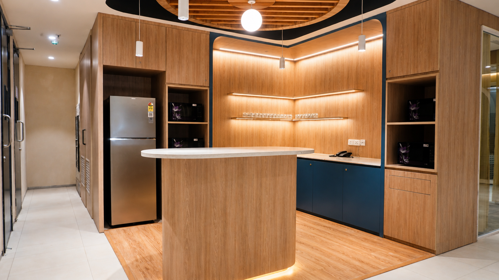 Modern office kitchenette with curved white island, wood cabinets, and warm backlit shelves.