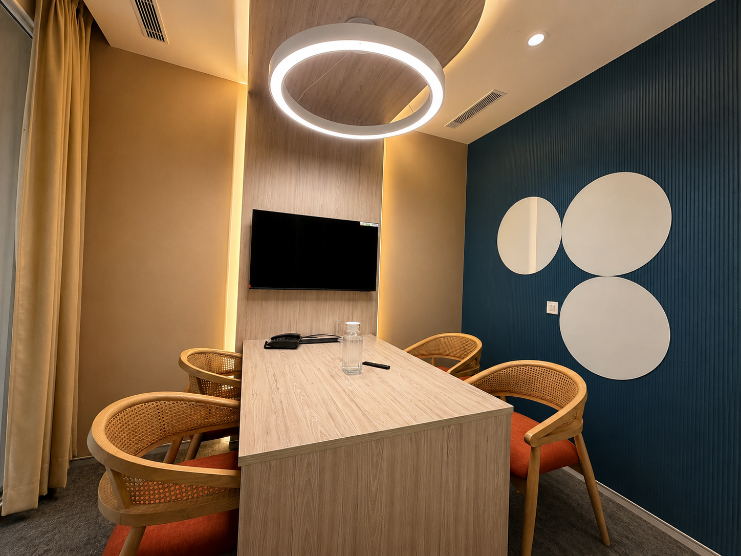 Modern meeting room with a long wooden table, circular ring light, wall-mounted TV, and woven chairs in beige and teal decor.